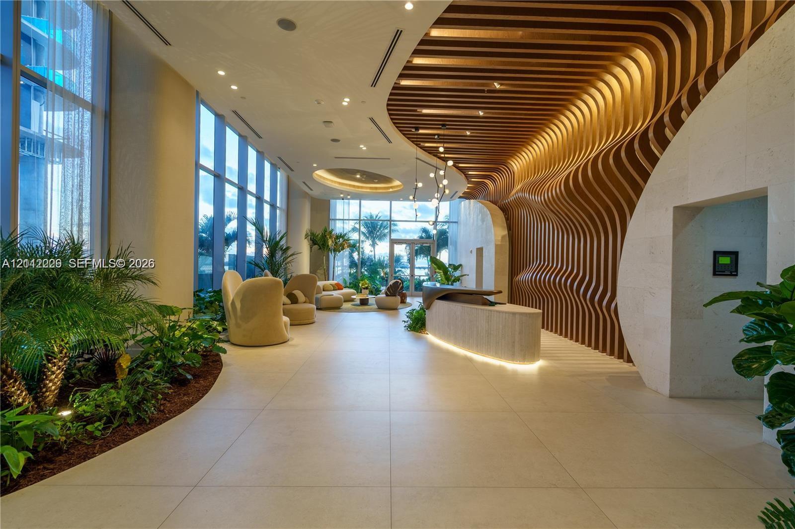 Grand lobby featuring floor-to-ceiling windows, custom organic-shaped wood slat wall detail, stone tile flooring, built-in reception desk with integrated lighting, and recessed ceiling elements