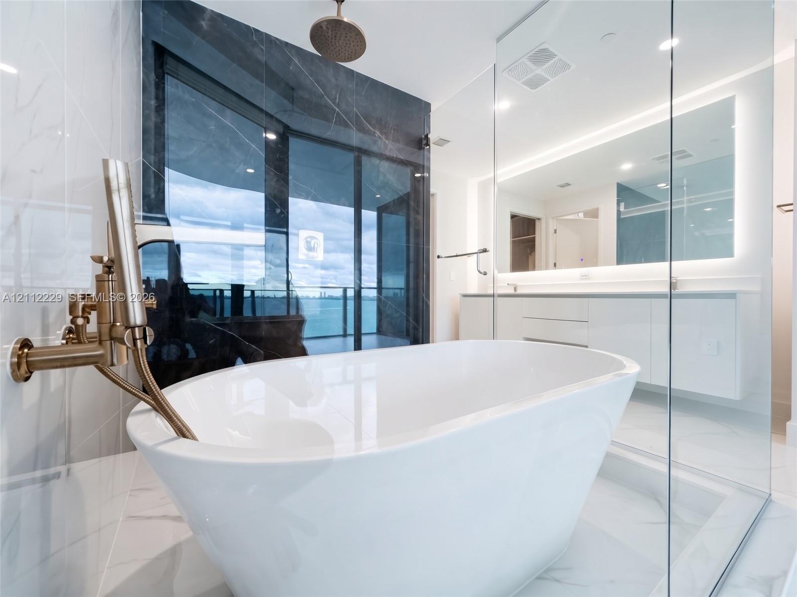 Freestanding soaking tub with brushed gold hardware, glass-enclosed shower, dual-vanity cabinetry, backlit wall mirror, and large-format tile flooring