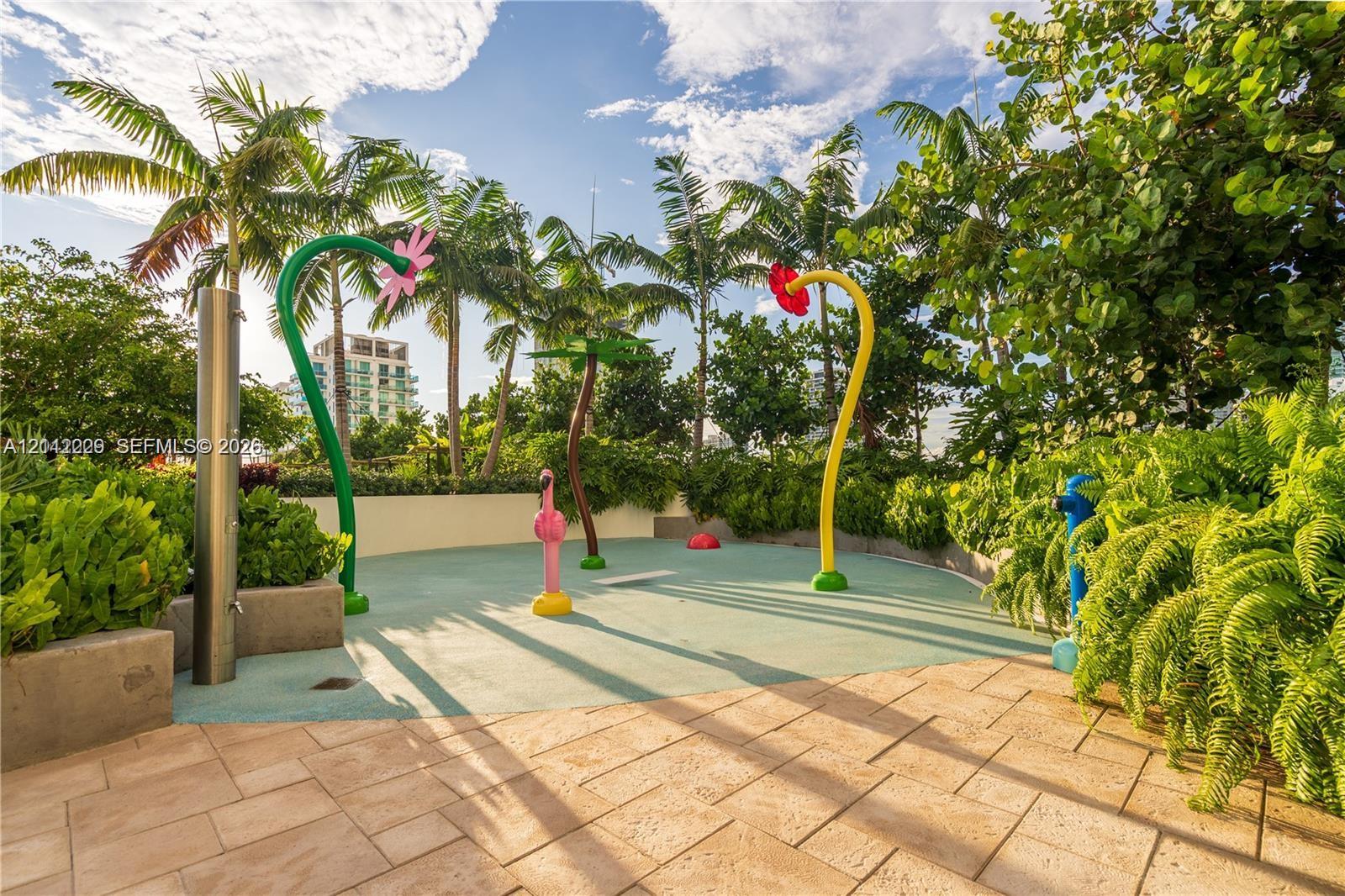 Outdoor splash pad area featuring aquatic play structures, textured soft-surface flooring, stone paver decking, and perimeter landscaping with mature palms