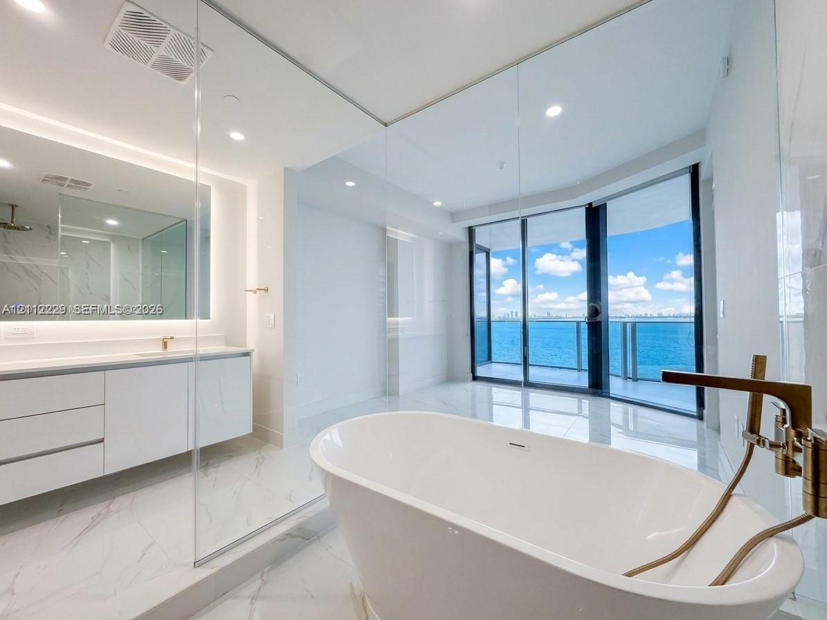 Freestanding soaking tub with a metallic faucet, white cabinetry featuring a light-toned countertop, glass-enclosed shower, large-format stone-finish floor tiling, and floor-to-ceiling glass sliding doors opening to a balcony
