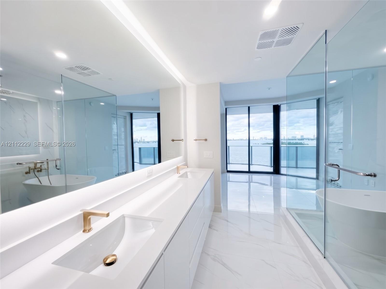 Dual-sink vanity with white countertops and gold-toned fixtures, floor-to-ceiling glass-enclosed shower, freestanding soaking tub, expansive wall-to-wall mirror with integrated lighting, and polished stone-finish flooring