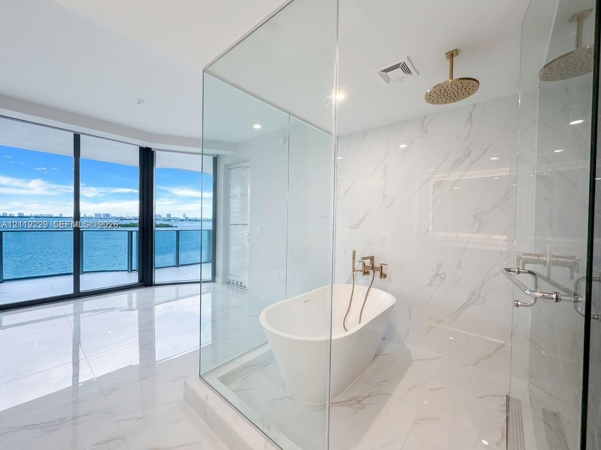 Floor-to-ceiling glass wall, freestanding soaking tub, rainfall shower head, stone-finish tile wall and flooring, and gold-tone fixtures