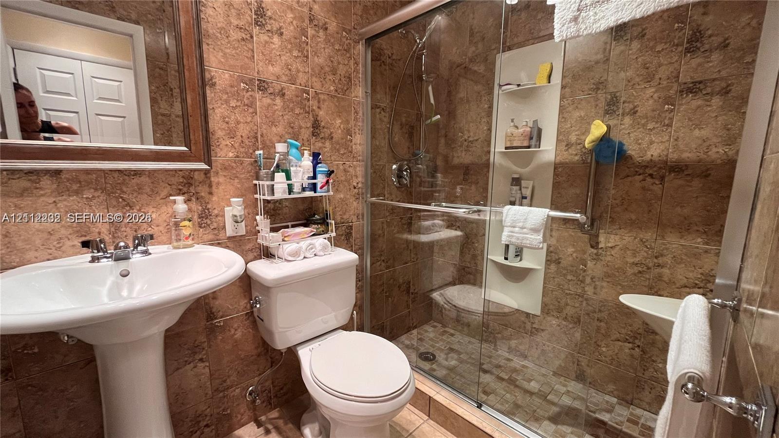 Bathroom featuring floor-to-ceiling stone wall tiling, a white pedestal sink with metallic fixtures, and a glass-enclosed shower with a built-in recessed shelf