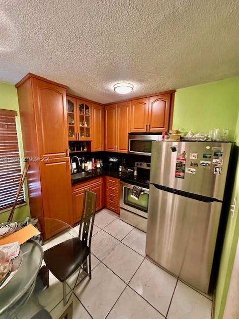 Kitchen featuring wood cabinetry with glass-panel upper doors, dark stone-finish countertops, stainless steel appliances, ceramic tile flooring, and a flush-mount ceiling light