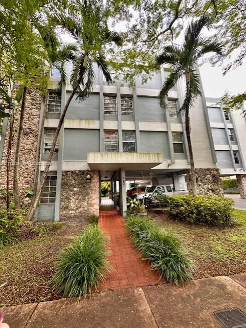 Multi-story facade featuring natural stone masonry, a covered entryway, and a brick-paved walkway surrounded by mature landscaping and palm trees