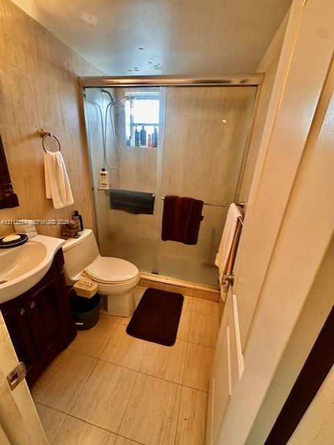 Bathroom featuring neutral stone-finish wall and floor tiling, dark wood cabinetry with a recessed sink, and a walk-in shower enclosure with a sliding glass door and integrated handheld showerhead