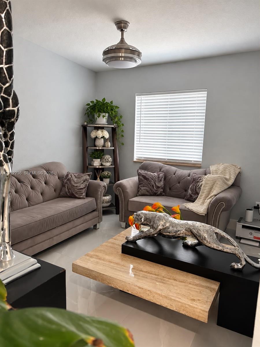 Neutral-toned interior space featuring polished tile flooring, a horizontal blind-covered window, and a brushed nickel ceiling fan with integrated lighting