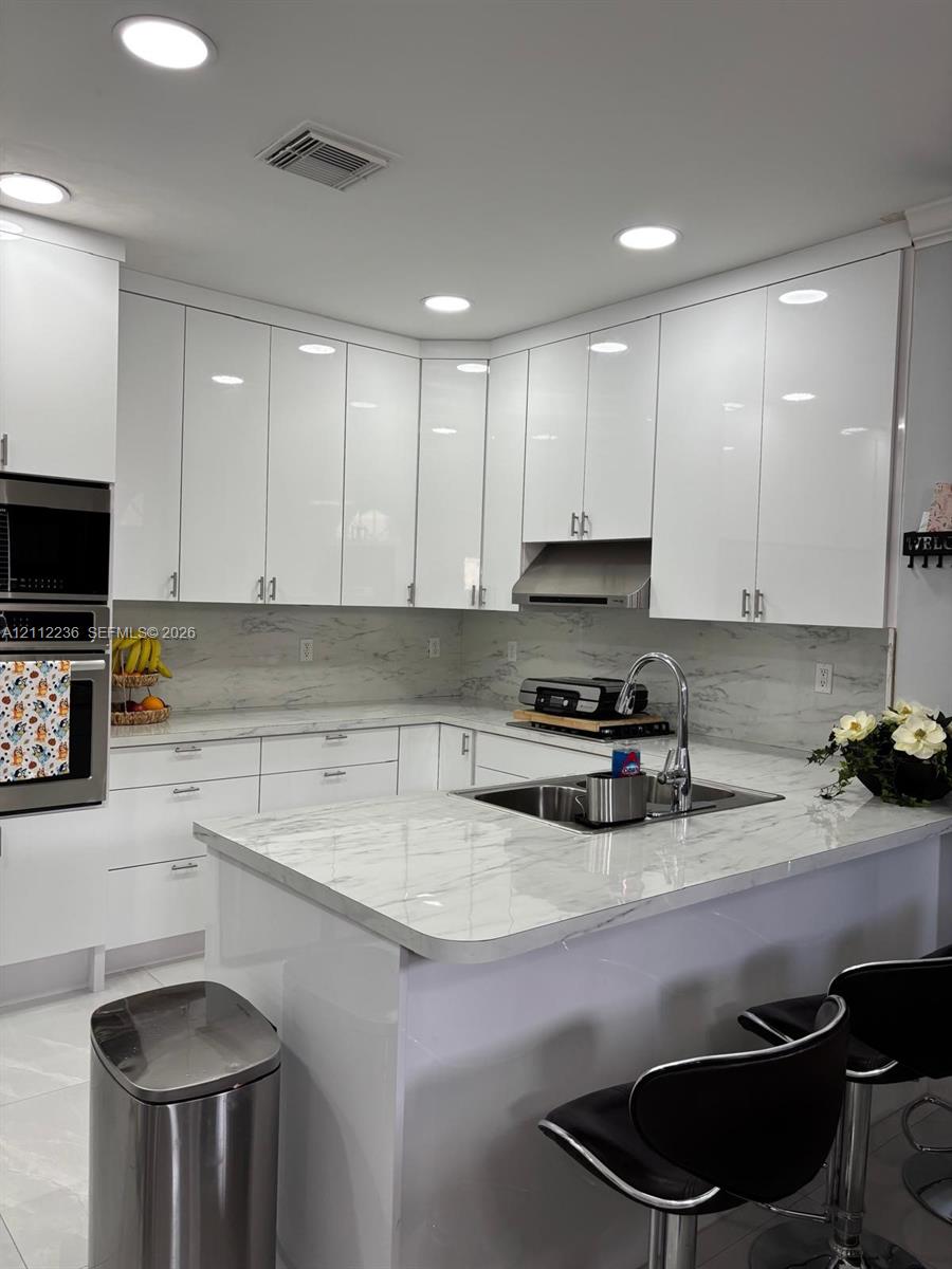 Kitchen featuring high-gloss white cabinetry, stone-patterned countertops, integrated stainless steel double oven, stainless steel range hood, and a double-basin undermount sink
