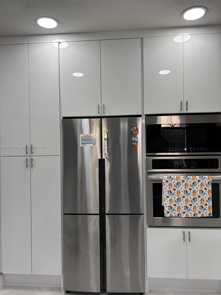 Floor-to-ceiling white high-gloss cabinetry, stainless steel French-door refrigerator, built-in stainless steel microwave, integrated stainless steel oven, and recessed ceiling lighting