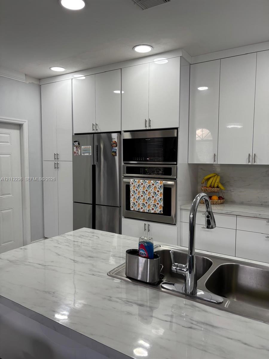 Kitchen featuring high-gloss white cabinetry, stone-finish countertops, stainless steel appliances, built-in double wall ovens, and a double-basin undermount sink with a gooseneck faucet