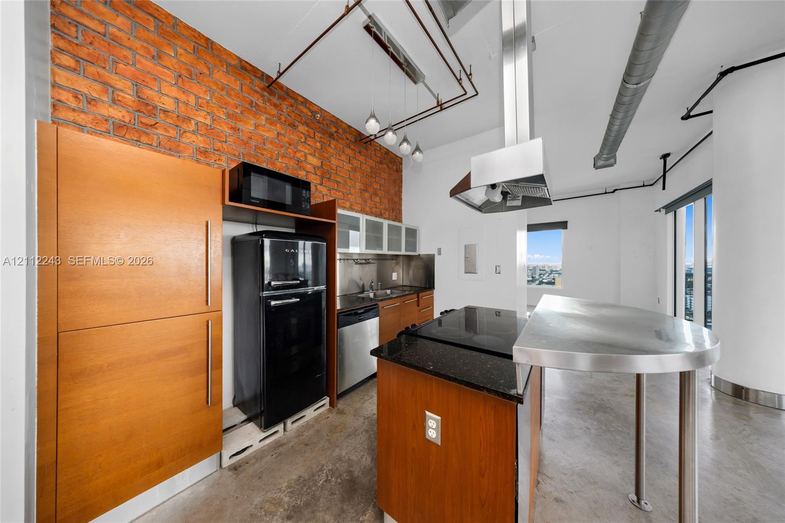 Kitchen featuring exposed brick wall, polished concrete flooring, stainless steel island with built-in cooktop, ceiling-mounted range hood, and wood-finish cabinetry