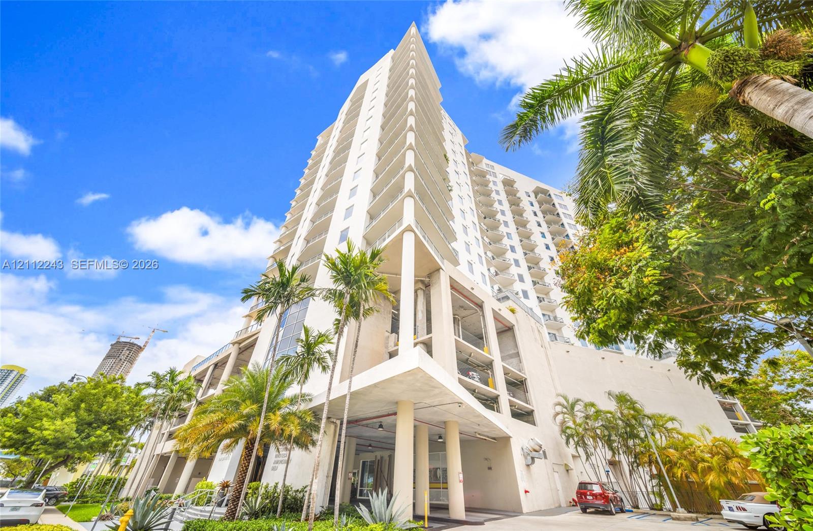 High-rise building exterior featuring tiered balconies, a structural concrete faade, and a covered ground-level arrival area with support pillars, accented by mature palm trees