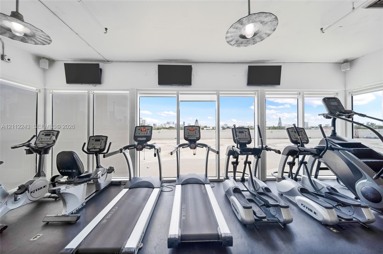 Fitness center featuring floor-to-ceiling windows, wall-mounted monitors, and industrial-style pendant lighting