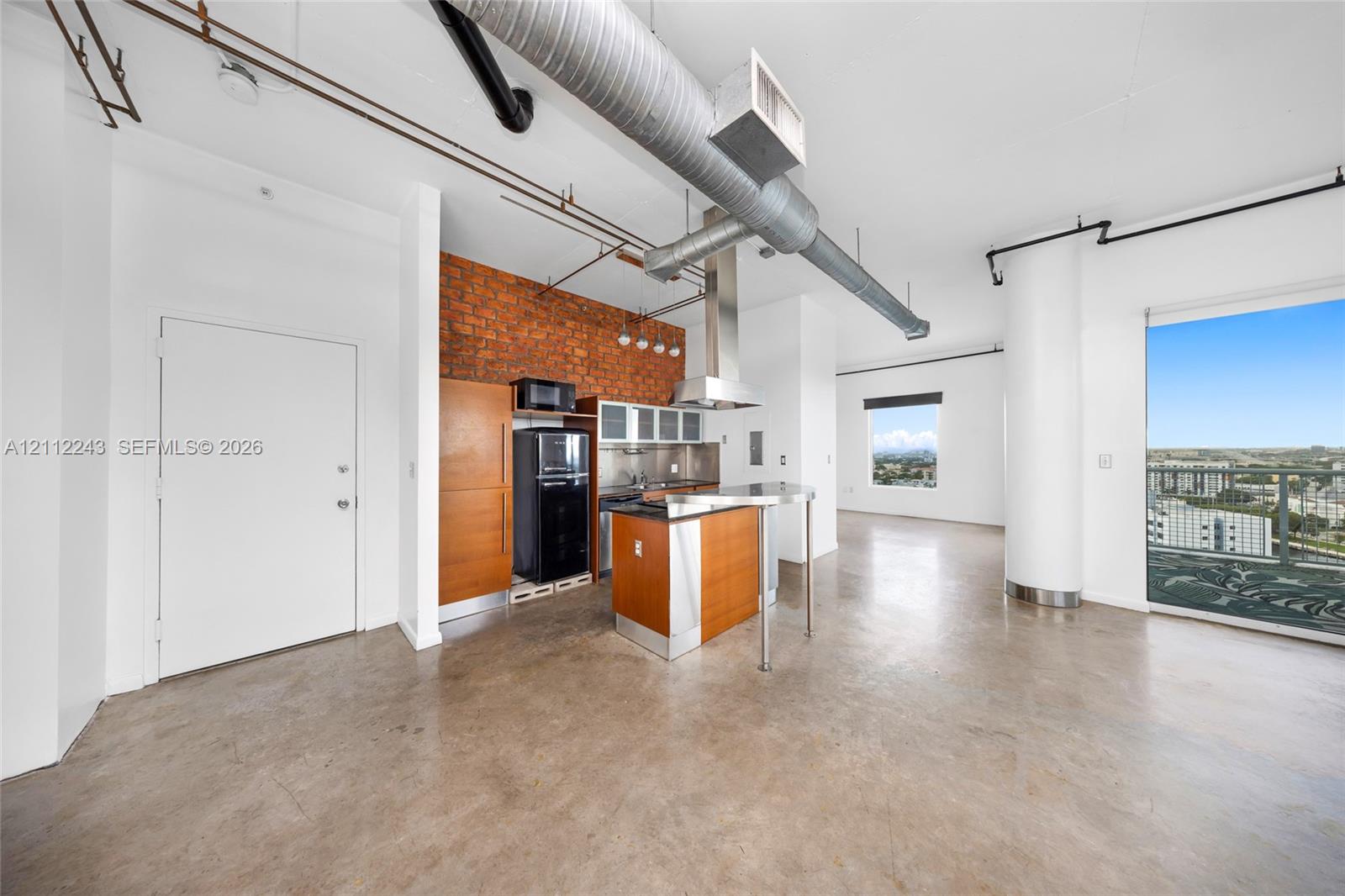 Industrial-style interior featuring polished concrete flooring, exposed brick accent wall, exposed ductwork, and floor-to-ceiling glass door providing exterior access