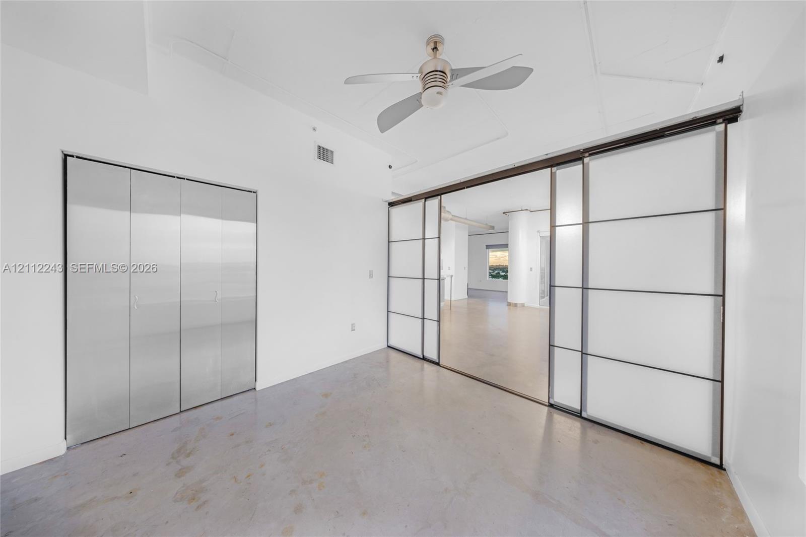 Interior space featuring polished concrete flooring, metallic folding closet doors, large frosted glass sliding panels with dark frames, and a ceiling-mounted fan