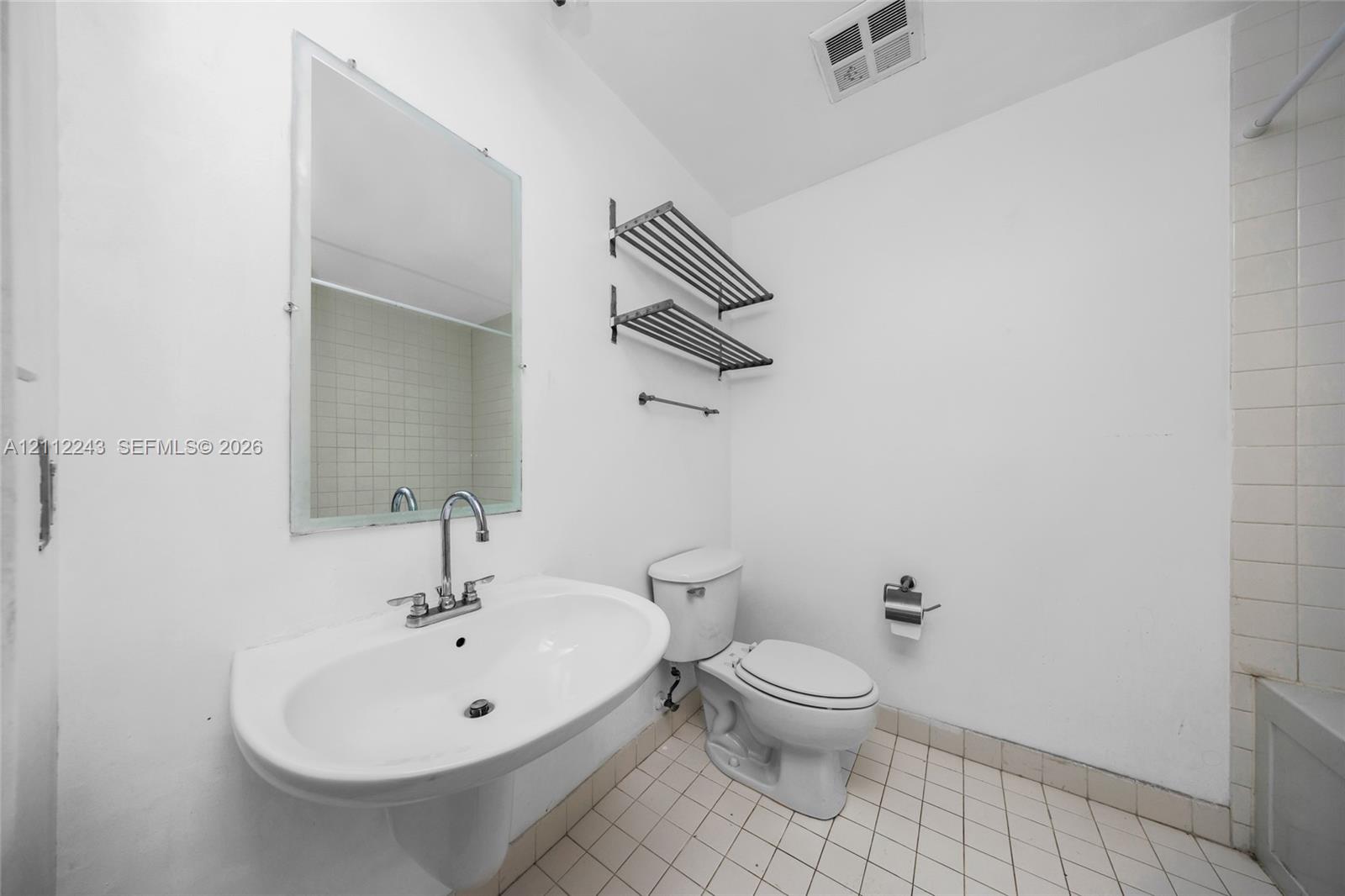 Wall-mounted ceramic pedestal sink with chrome faucet, frameless rectangular mirror, dual metal wall-mounted shelving, and tiled flooring