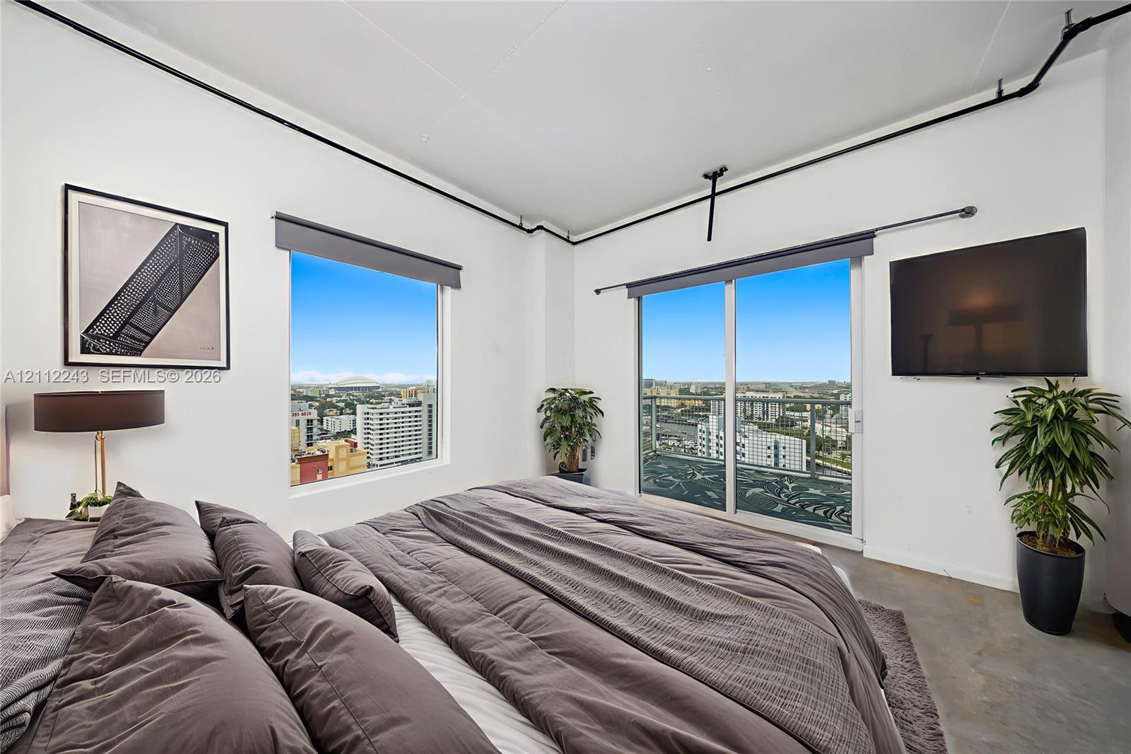 Industrial-style room featuring polished concrete flooring, exposed ceiling-mounted piping, sliding glass door to a private balcony, and large windows with urban exterior scenery