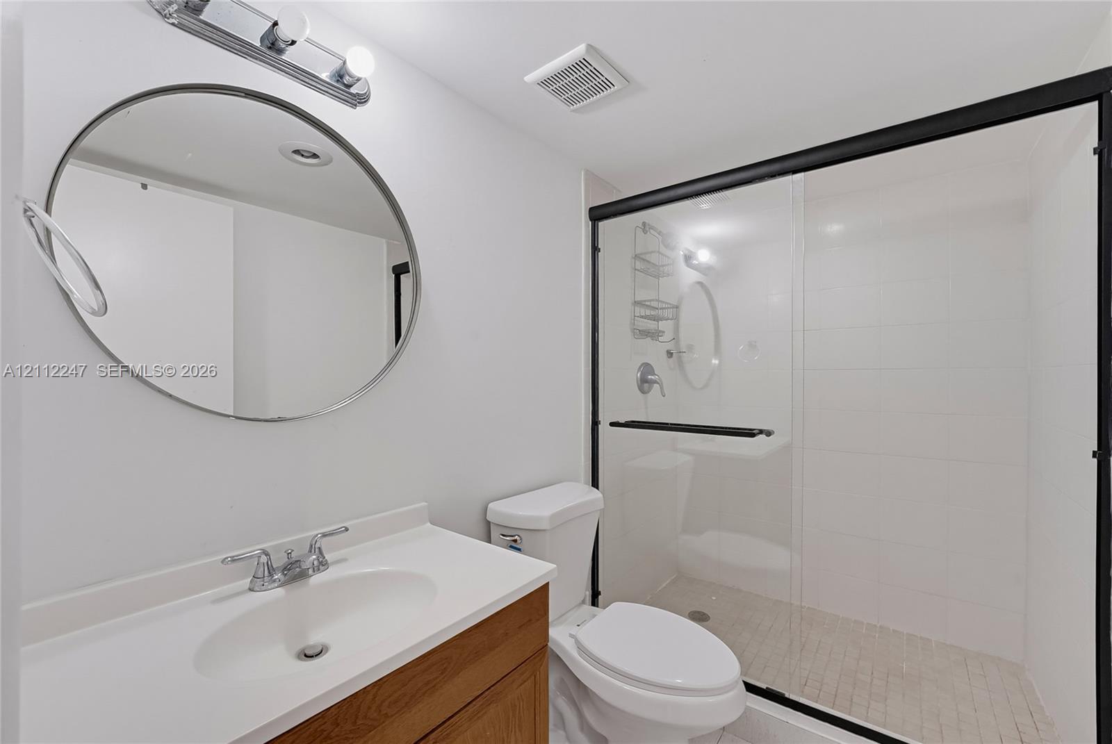 Bathroom featuring a wood-finish vanity, round mirror with metallic frame, wall-mounted light fixture, and a glass-enclosed shower with tiled walls