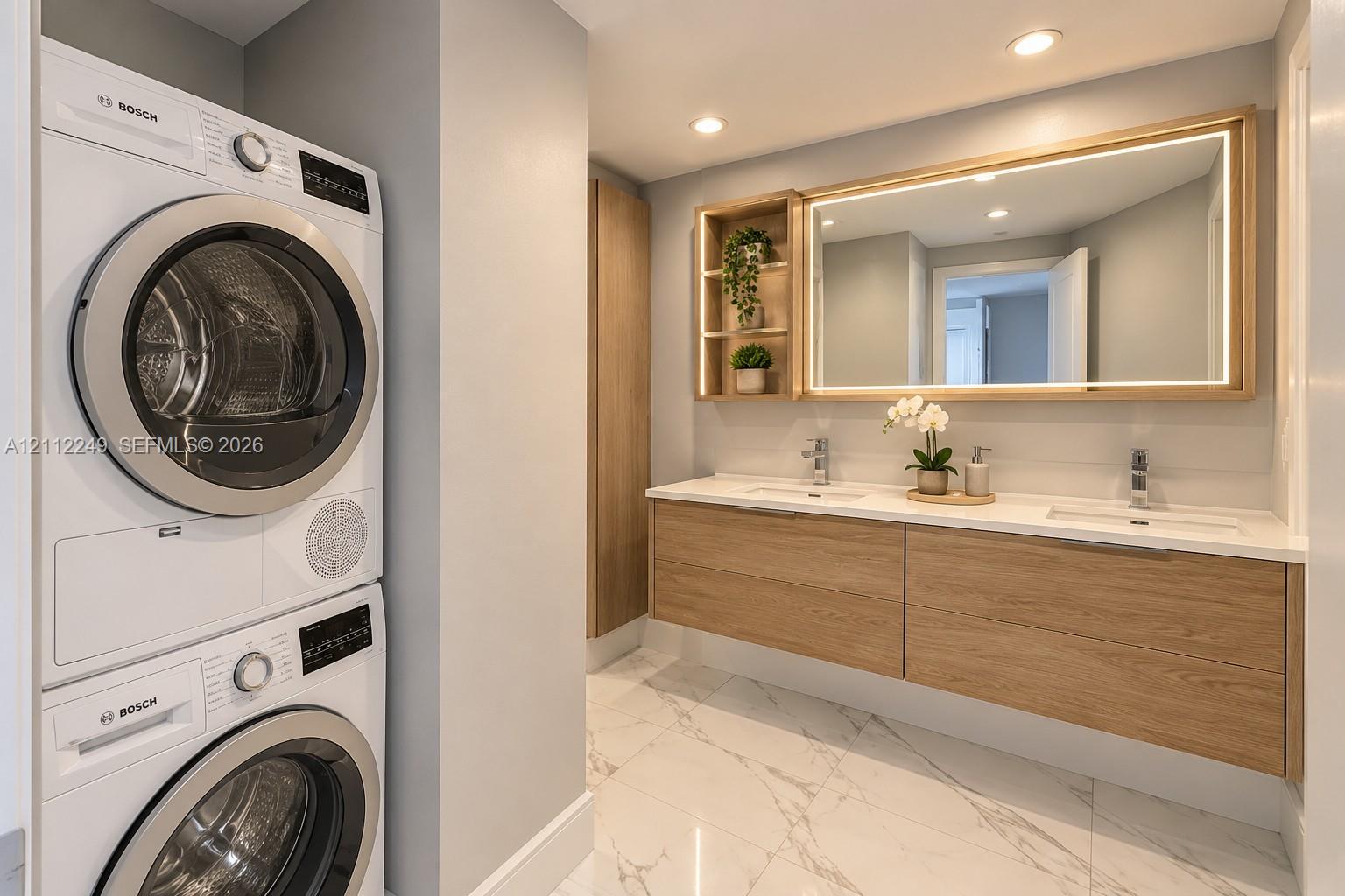 Floating wood-finish vanity with dual integrated sinks, stone-finish countertop, LED-backlit mirror, built-in shelving, and stacked Bosch laundry appliances on polished tile flooring
