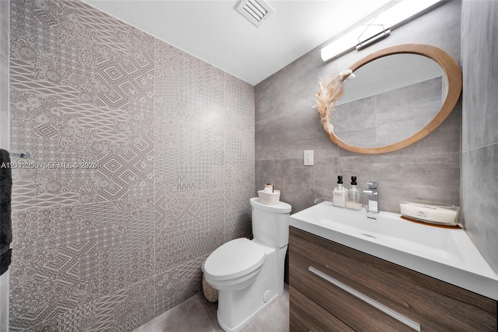 Powder room featuring a wood-finish vanity with an integrated basin, large-format grey tile walls, a decorative patterned accent wall, and an oval mirror with a natural wood frame