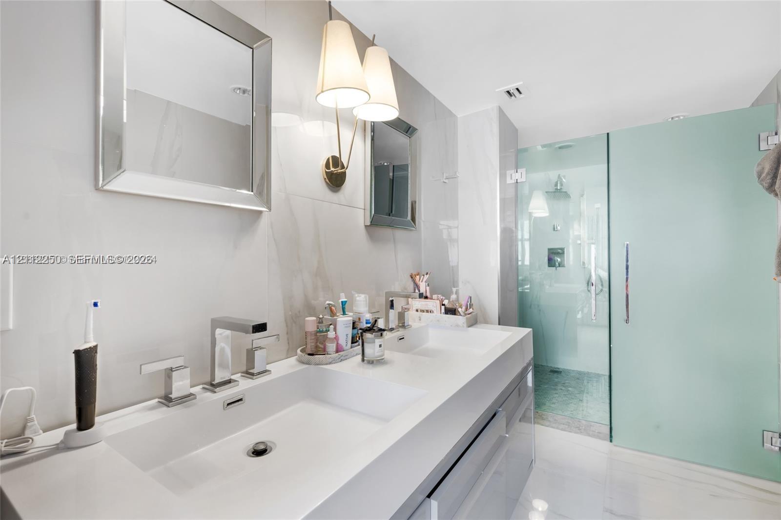 Dual-sink vanity with a solid-surface countertop, rectangular basins, wall-mounted polished chrome faucets, and frameless glass shower enclosure