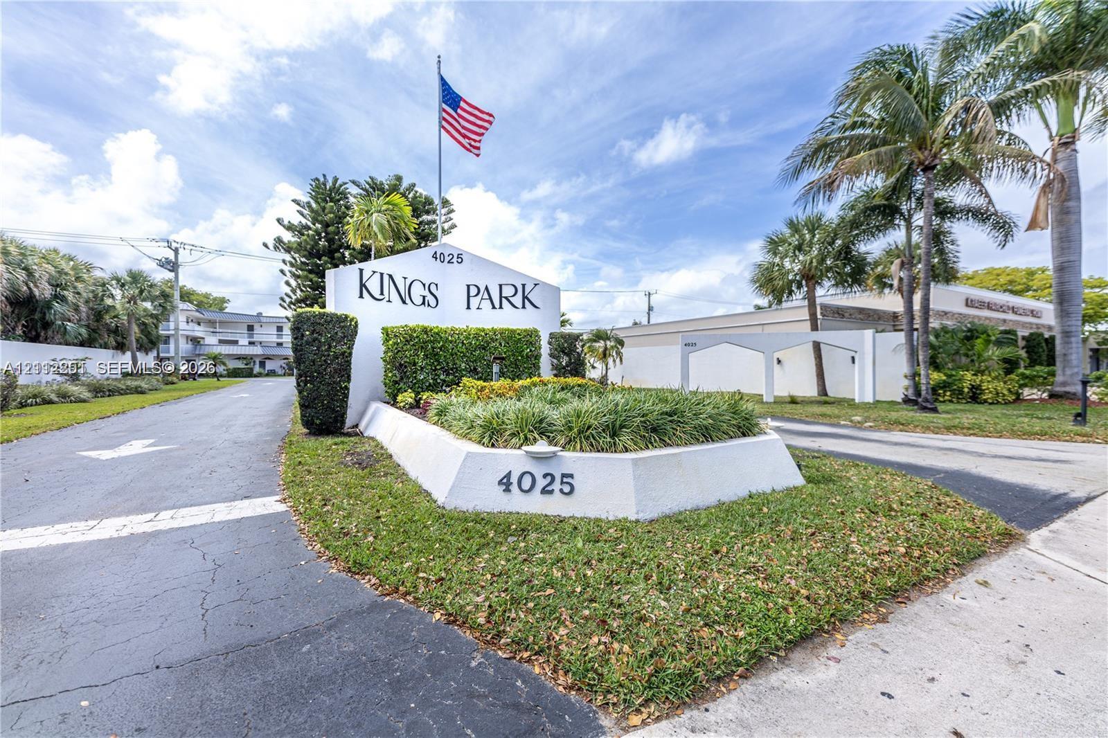 Kings Park entrance featuring a stucco monument sign, manicured landscaping, asphalt driveway, and mature palm trees