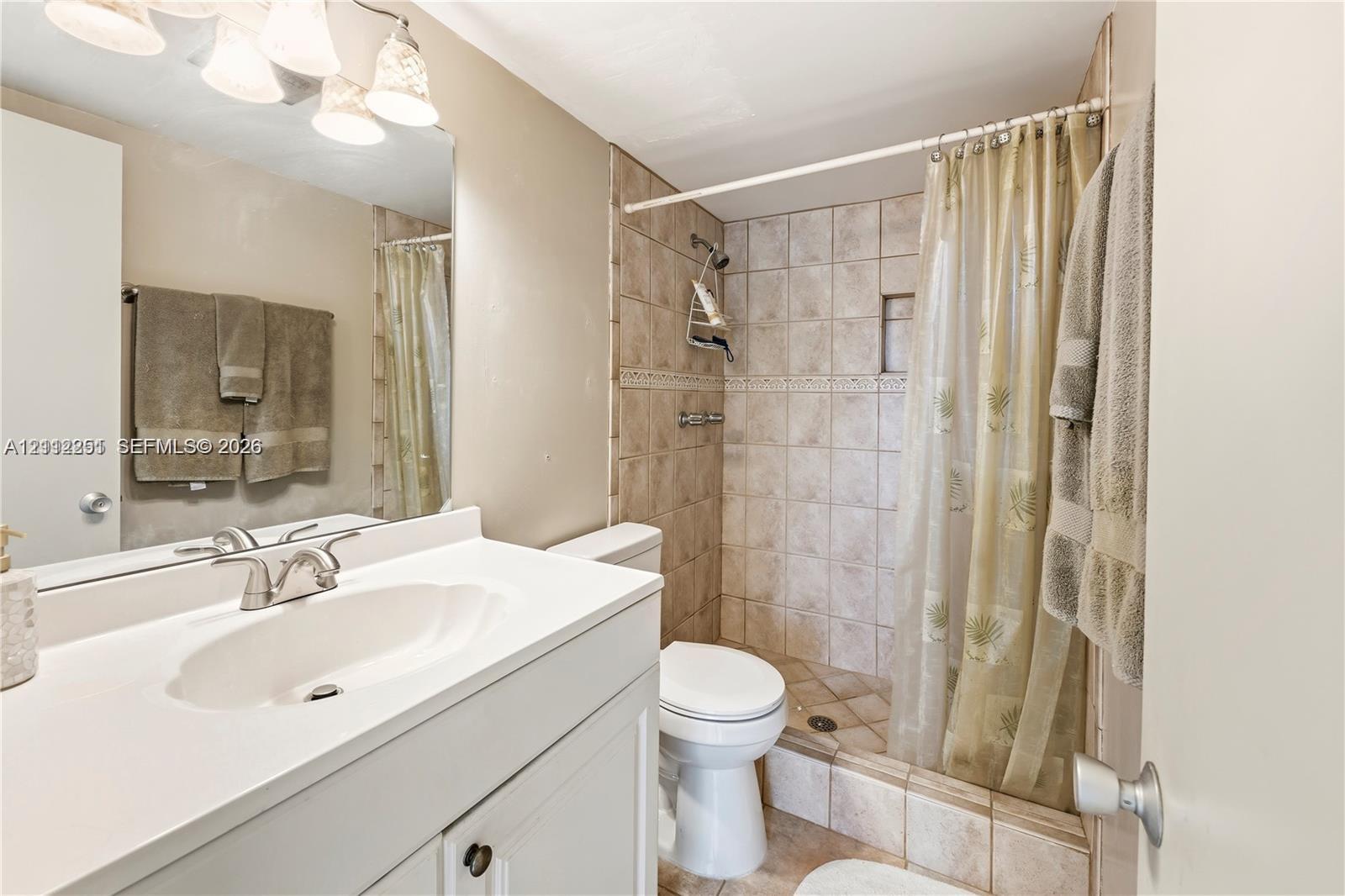 Bathroom featuring a white vanity with an integrated countertop, polished chrome faucet, full-width mirror, light fixture with four frosted glass shades, and a tiled shower stall with a decorative accent border