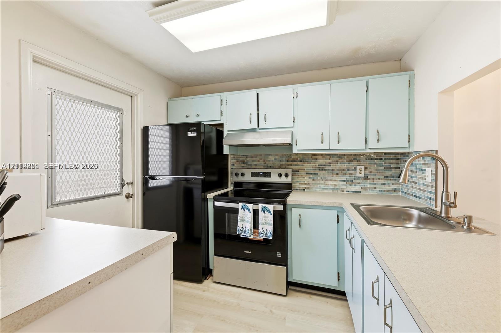Kitchen featuring pale blue cabinetry, solid surface countertops, multi-toned mosaic tile backsplash, stainless steel range with overhead hood, and light-tone wood-finish flooring Kitchen featuring pale blue cabinetry, solid surface countertops, multi-toned mosaic tile backsplash, stainless steel range with overhead hood, and light-tone wood-finish flooring
