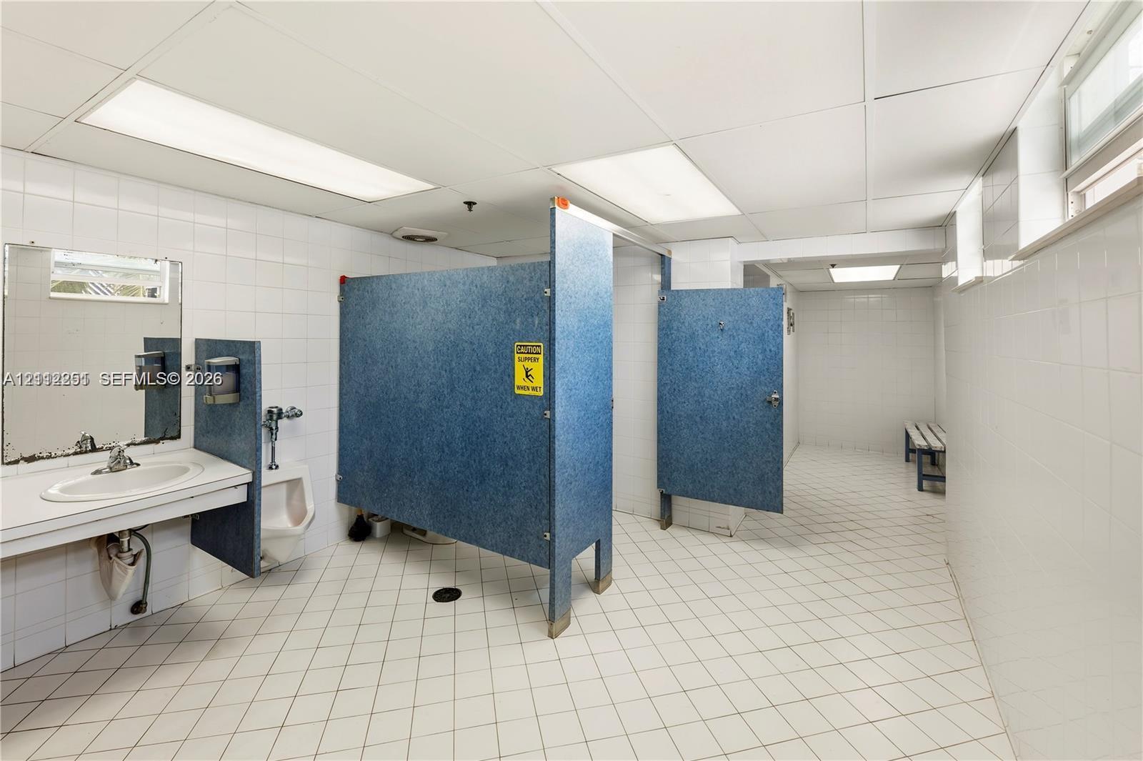 Restroom featuring ceramic tile walls and flooring, drop ceiling with recessed lighting, blue privacy partitions, wall-mounted sink, and built-in bench seating