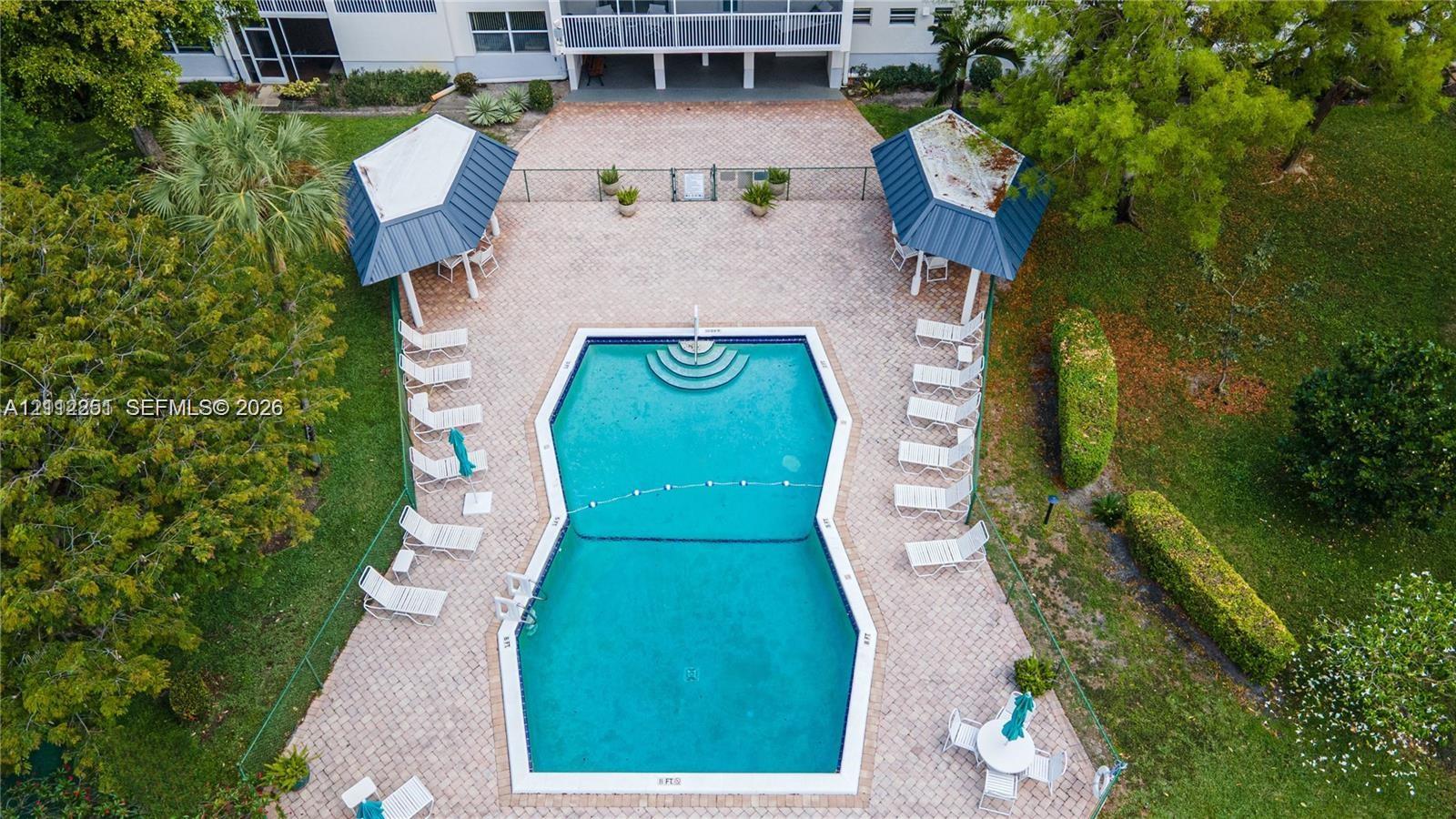 Swimming pool with built-in stairs, surrounded by a paved deck, two covered cabanas with metal roofing, and landscaped grounds