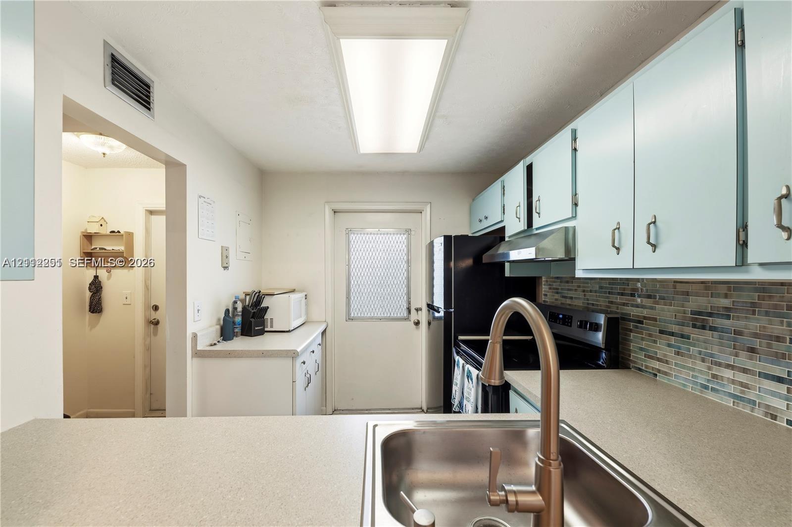 Kitchen featuring a stainless steel undermount sink, gooseneck faucet, solid-surface countertops, multicolored glass tile backsplash, and recessed panel cabinetry