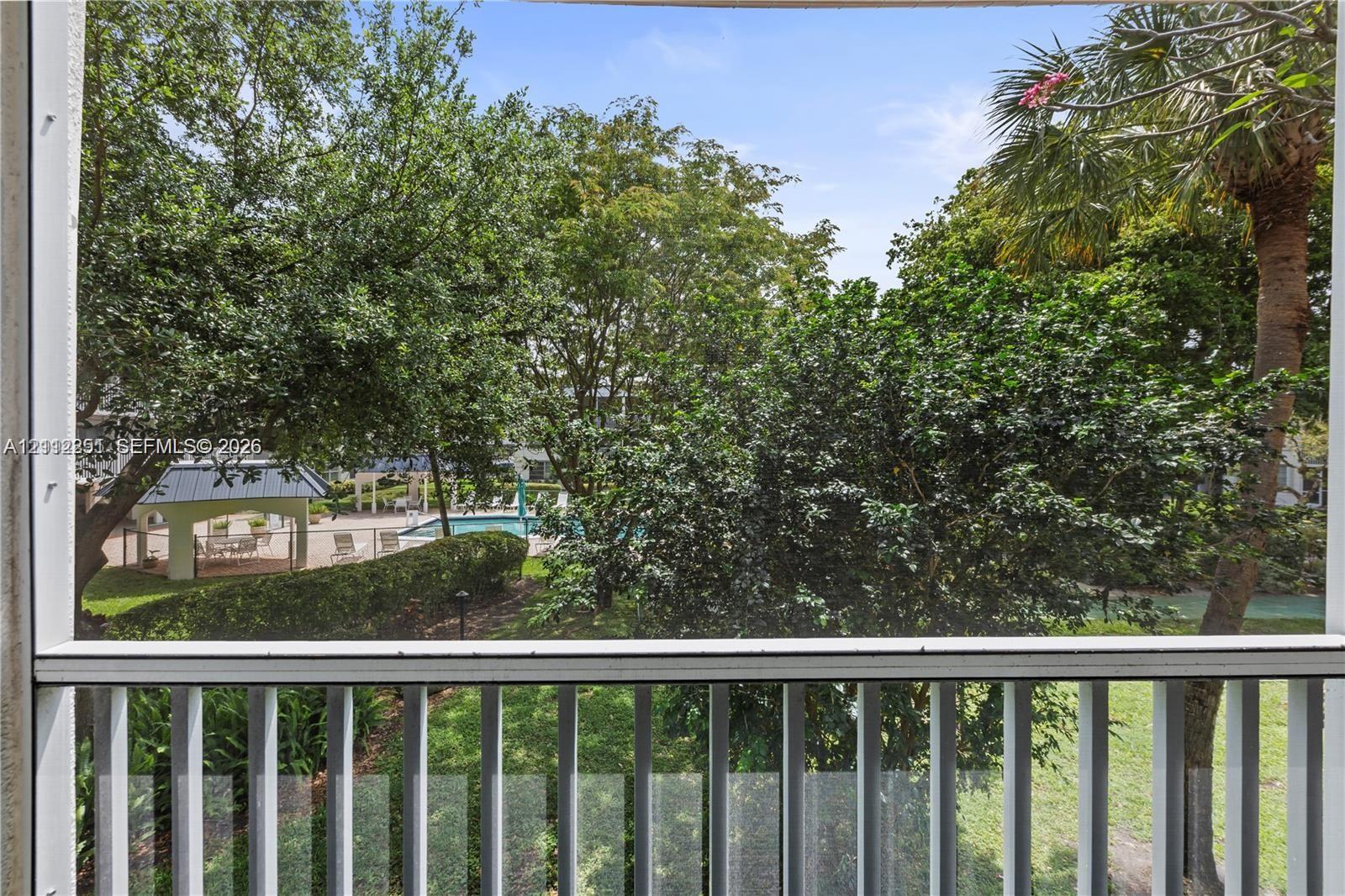 Screened-in balcony with white metal railings, overlooking a swimming pool area, manicured landscaping, mature trees, and a pavilion structure