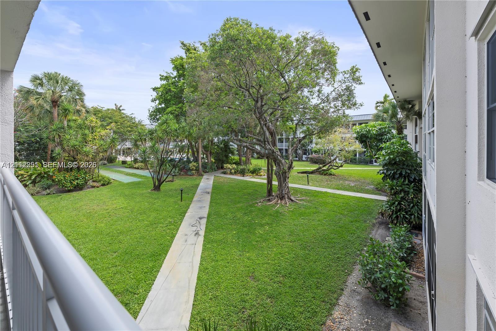 Covered balcony with white railing overlooking a landscaped common area featuring mature trees, manicured lawn, and concrete walkways