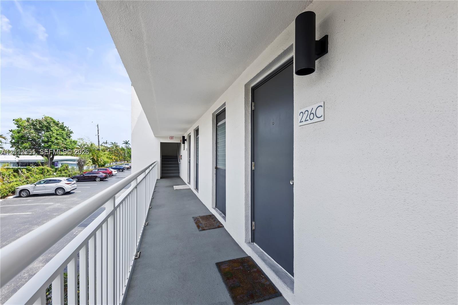 Exterior walkway featuring white stucco walls, grey entry doors, vertical matte black sconce lighting, and a white metal railing