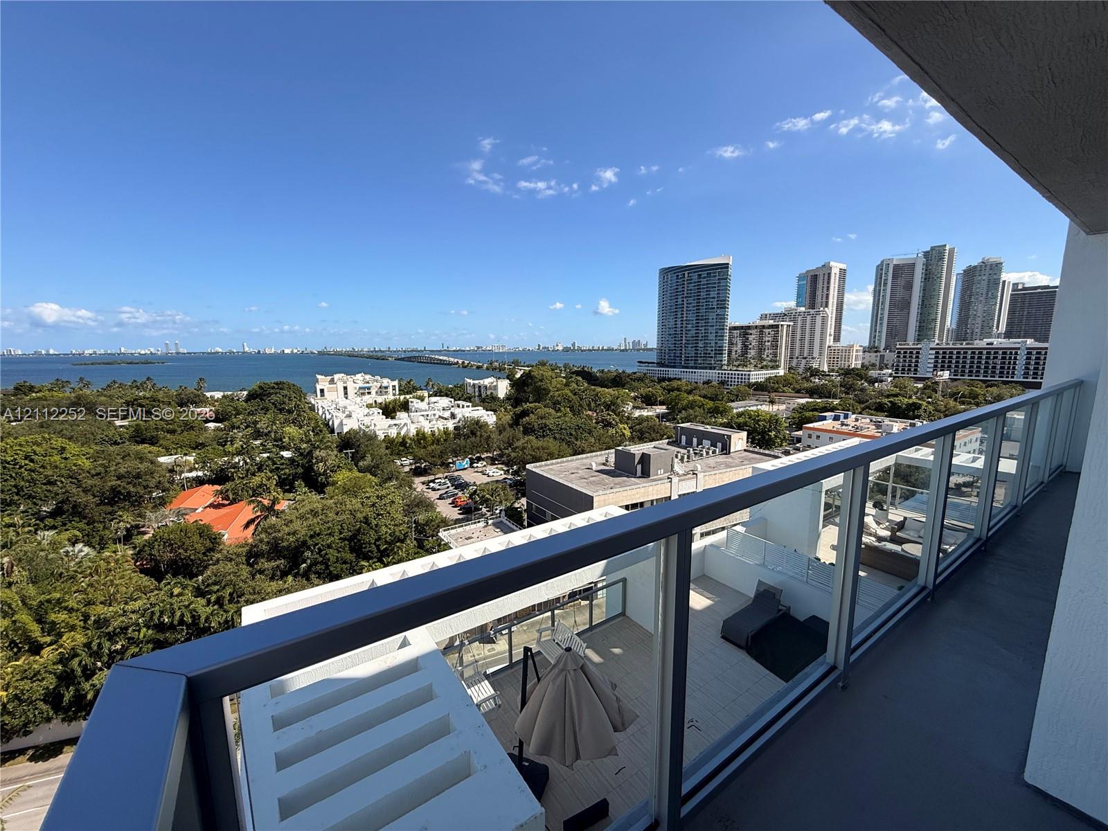 Covered balcony featuring glass railing panels and metal handrails, overlooking a coastal landscape with high-rise structures and open water