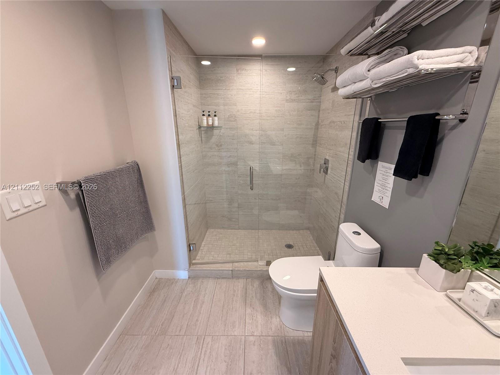 Walk-in shower with frameless glass enclosure, floor-to-ceiling stone-style wall tile, built-in shower niche, light-toned stone-style floor tile, and vanity with solid-surface countertop