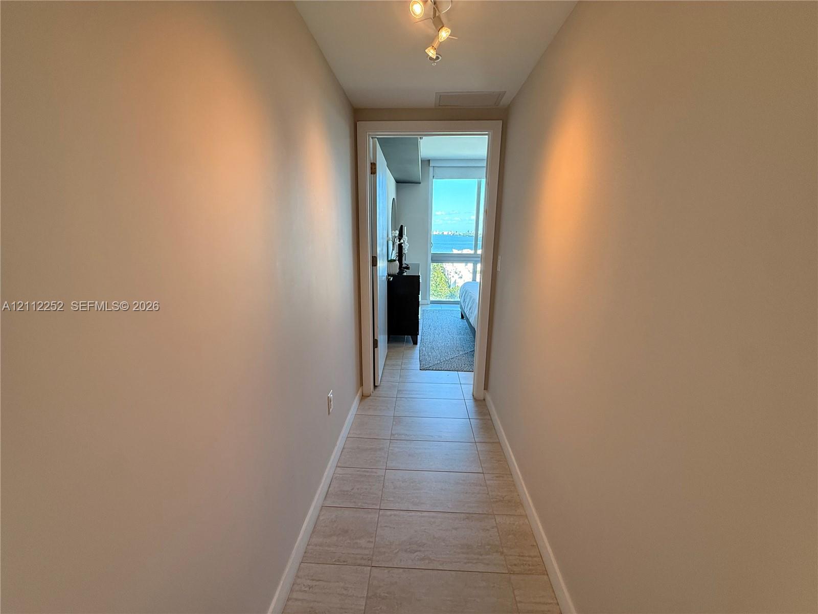 Hallway featuring neutral wall tones, tile flooring, and multi-directional track lighting, leading toward an exterior window
