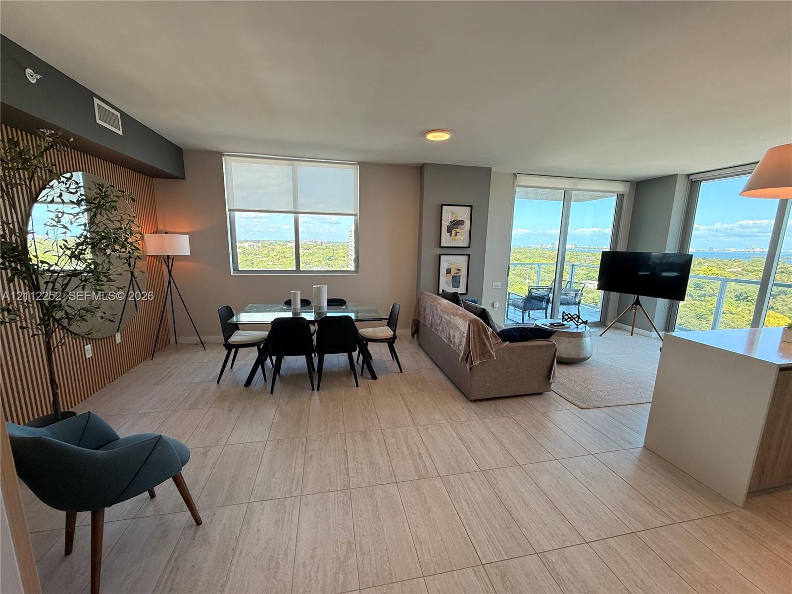 Open-concept living area featuring neutral tile flooring, floor-to-ceiling windows, natural wood-slat accent wall, and glass sliding doors providing access to a private balcony