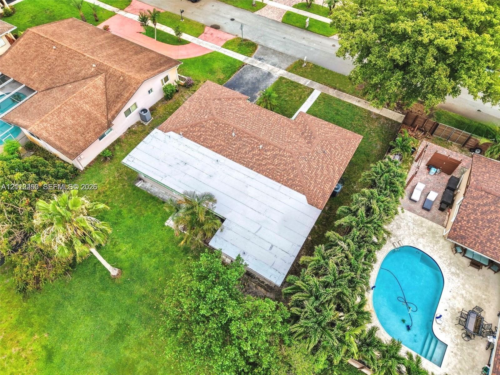 Aerial perspective featuring a single-story residence with a shingled roof, a paved patio, an in-ground swimming pool, and mature landscaping