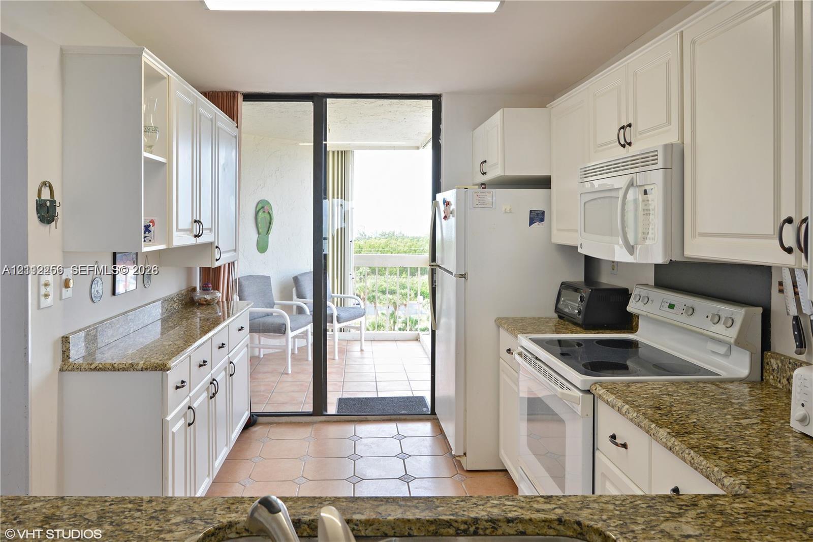 Kitchen featuring white shaker-style cabinetry, stone-finish countertops, tile flooring, and integrated white appliances, with sliding glass doors opening to a tiled balcony
