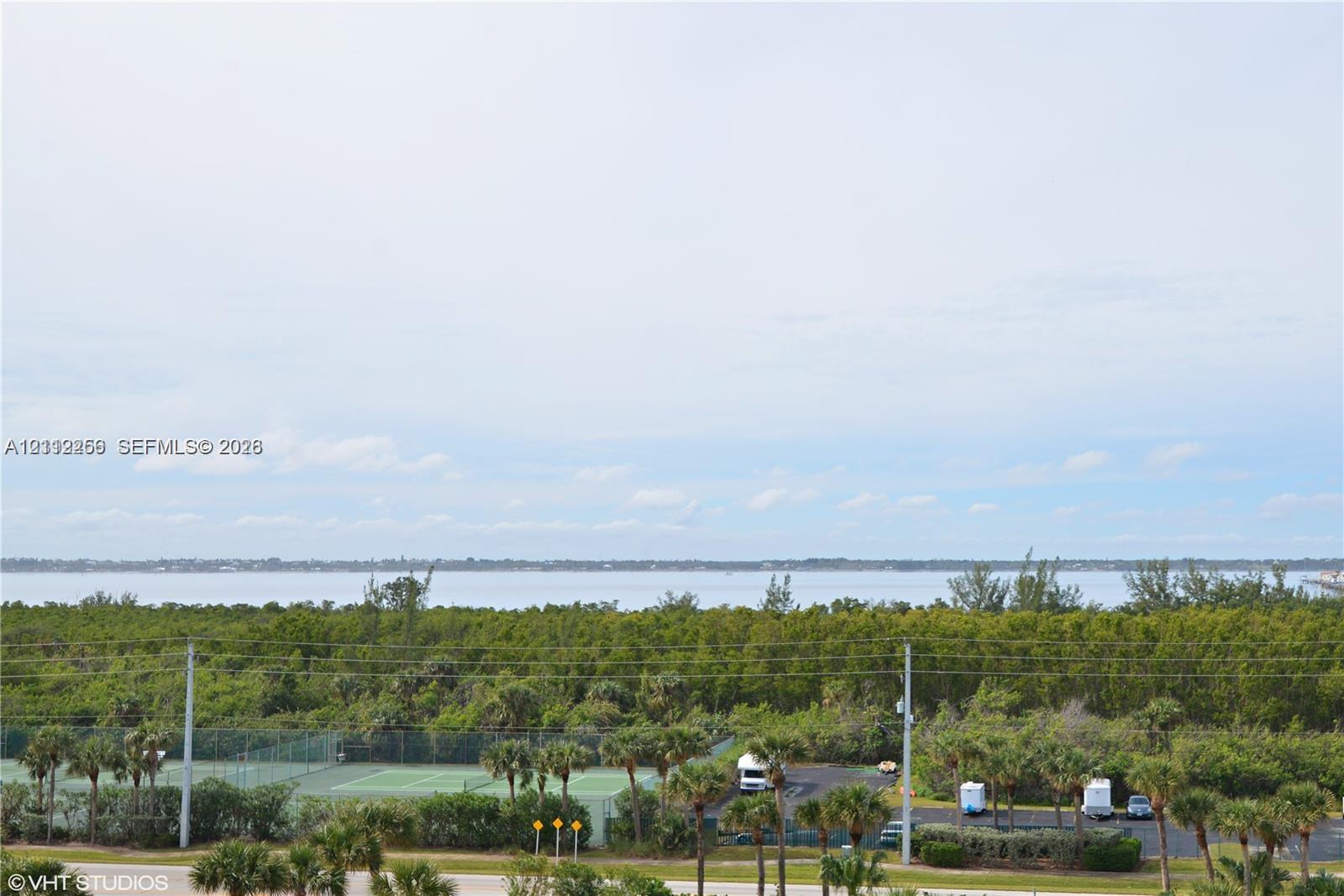 Expansive water horizon with mature treeline, featuring an outdoor tennis court and paved parking area