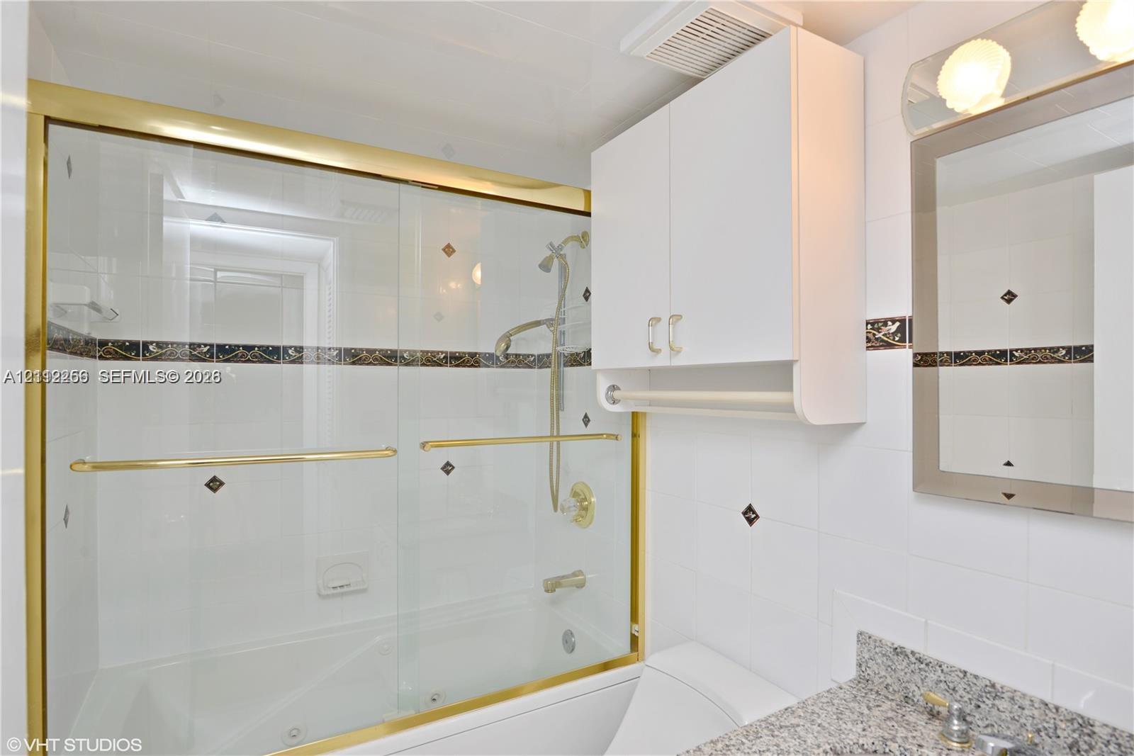 Bathroom featuring a tub-shower combination with sliding glass doors, gold-tone metal trim, stone-composite vanity countertop, wall-mounted white cabinetry, and decorative tile accents