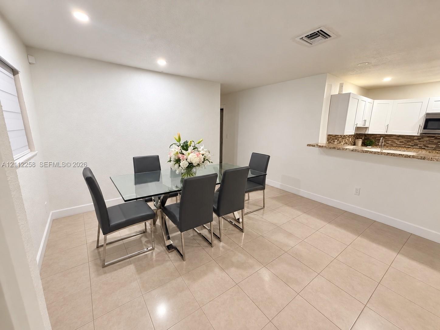 Open-concept space featuring large-format neutral floor tile, white shaker-style cabinetry, a stone-patterned backsplash, and a breakfast bar Open-concept space featuring large-format neutral floor tile, white shaker-style cabinetry, a stone-patterned backsplash, and a breakfast bar