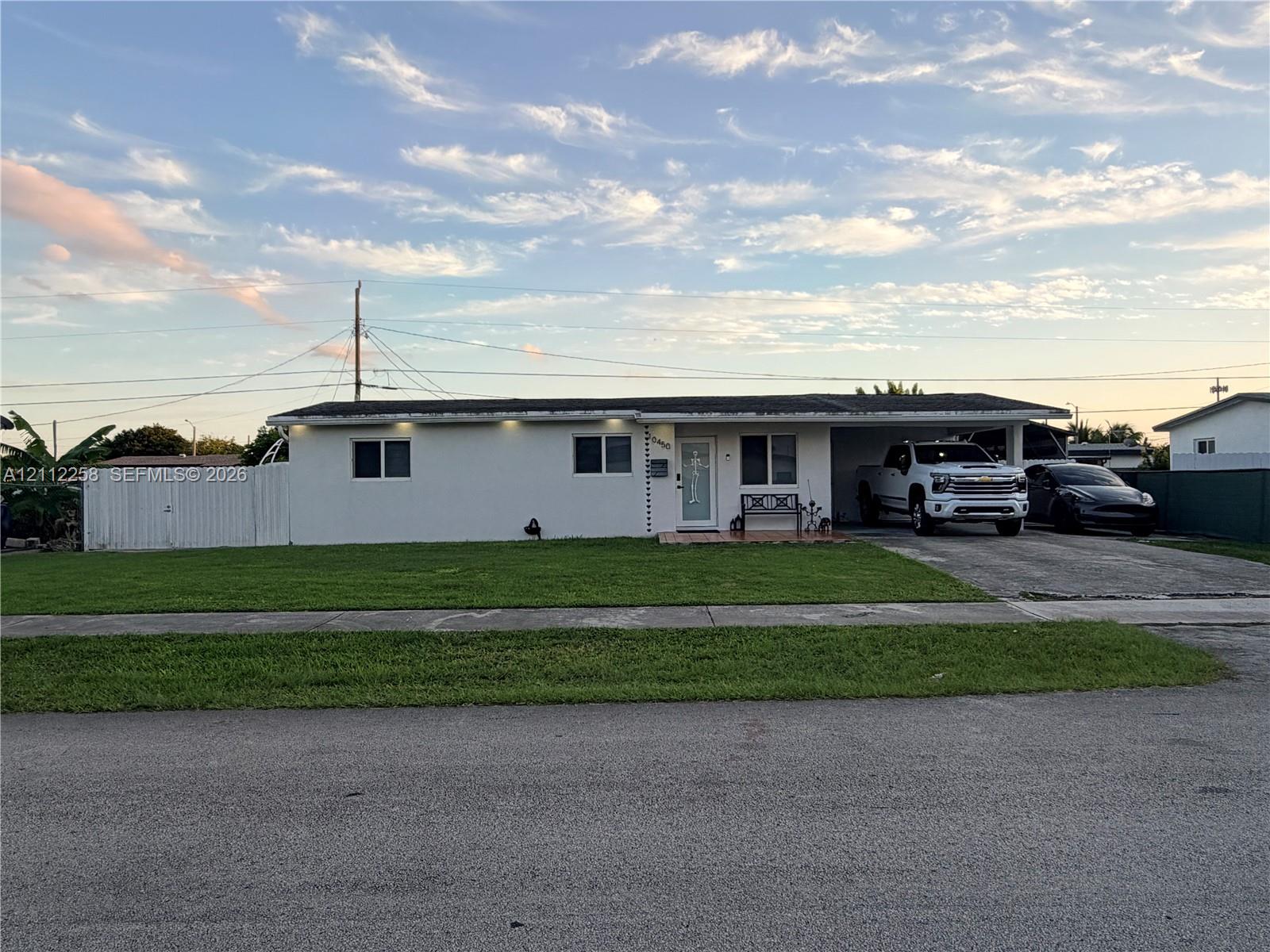 Single-story exterior featuring white stucco siding, a covered carport, recessed exterior lighting, and a white privacy fence