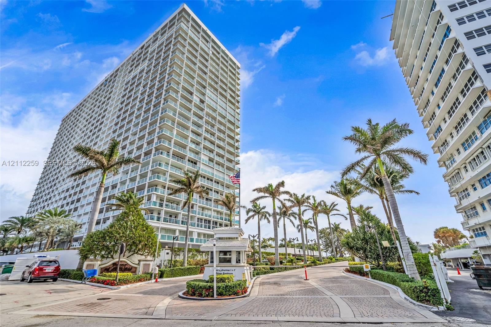 High-rise exterior featuring private balconies with glass railings, a paved motor court, a guardhouse entry, and mature palm landscaping