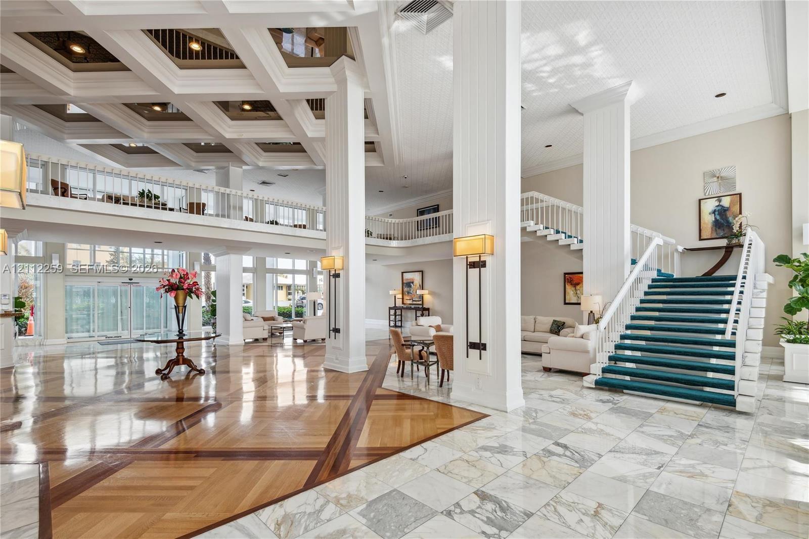 Grand entrance featuring high coffered ceilings, stone-tile flooring with wood-finish inlays, decorative white columns, and a double-railed staircase Grand entrance featuring high coffered ceilings, stone-tile flooring with wood-finish inlays, decorative white columns, and a double-railed staircase