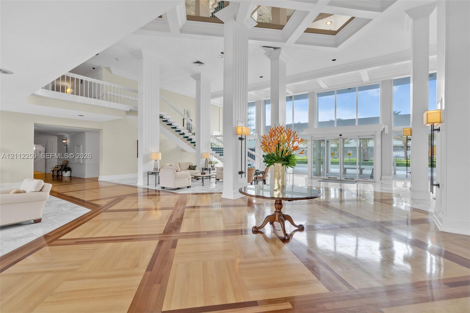Grand atrium featuring a coffered ceiling, polished wood-finish flooring with intricate inlay, soaring structural columns, expansive floor-to-ceiling glass entry doors, and a decorative staircase