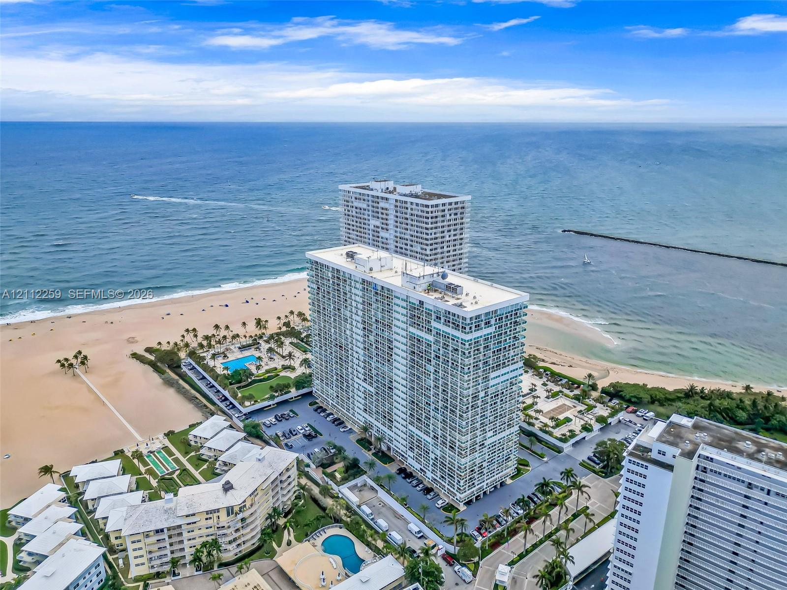 Aerial perspective of a high-rise residential complex featuring direct beach access, a swimming pool, paved surface parking, and coastal landscape