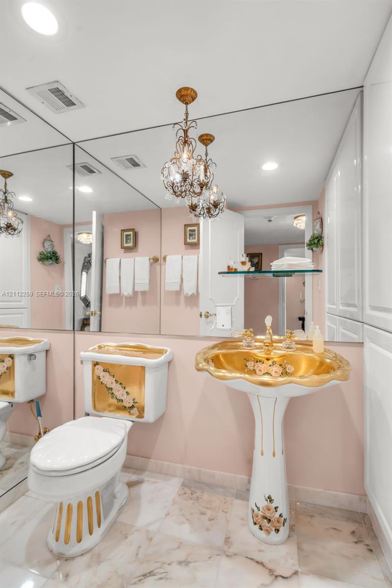 Bathroom featuring a floor-to-ceiling mirror, pedestal sink with gold-tone accents and floral detailing, matching commode, and polished stone flooring with decorative inlay