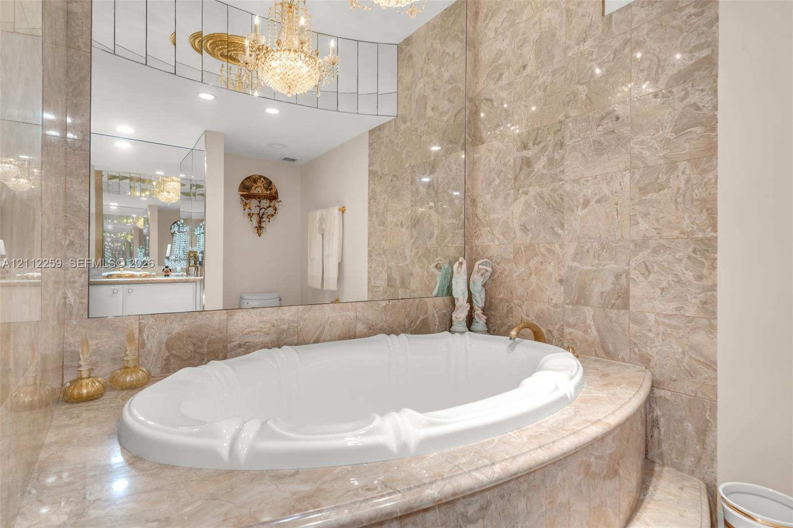 Oval soaking tub featuring a decorative basin, surrounded by large-format natural stone tiling with an expansive wall-to-wall mirror, crystal-style chandelier, and recessed lighting